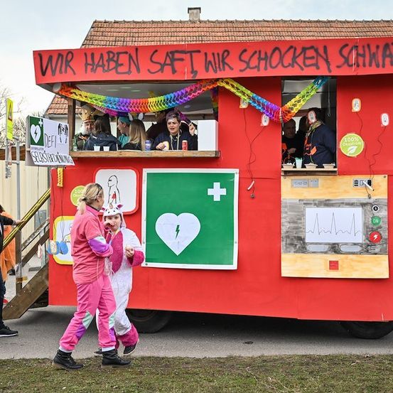 Eine Gruppe von Menschen in pinken und weißen Kostümen geht an einem roten Imbisswagen mit Herzsymbol und einem Schild vorbei, das 'Wir haben Saft, Wir schocken Schwandorf' besagt.