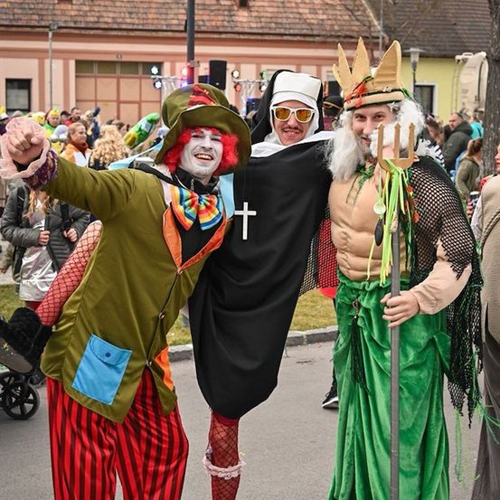 Drei Menschen in bunten Kostümen posieren für ein Foto auf einer Straße während eines Festivals. Einer trägt ein Clownkostüm, ein anderer ein Nonnenkostüm und der dritte ein Dreizack-haltendes Kostüm.