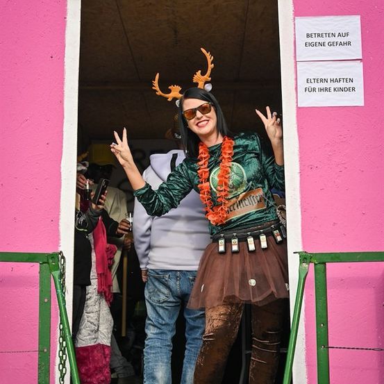 Eine Frau in einem grünen Kostüm mit Hirschgeweih und Sonnenbrille steht in einer pinken Kabine mit einem Schild, das 'Auf eigene Gefahr betreten' und einem weiteren Schild, das 'Eltern, achten Sie auf Ihre Kinder' sagt.