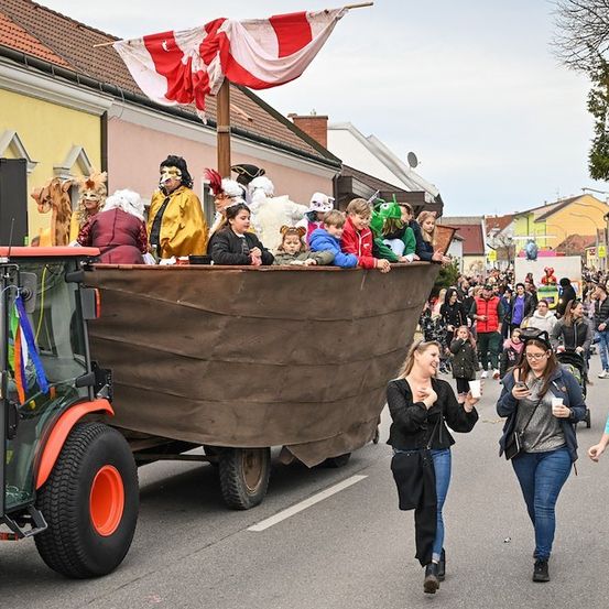 Ein großer Wagen mit kostümierten Personen wird von einem Traktor gezogen, während eine Parade stattfindet, und eine Menge von Zuschauern beobachtet.