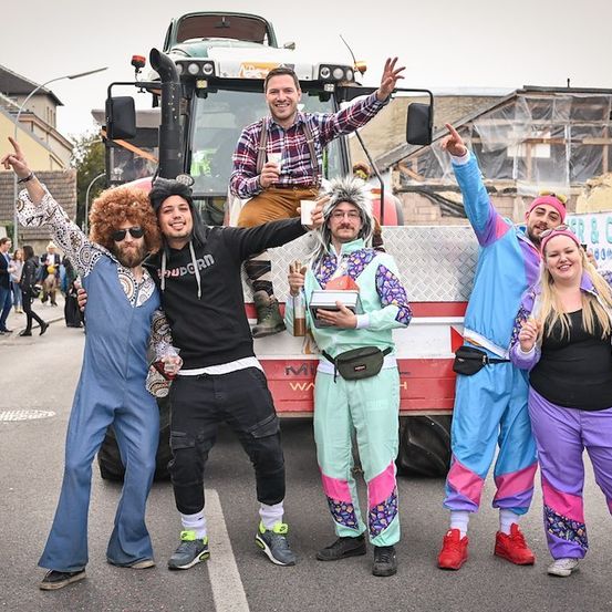 Eine Gruppe von Menschen in bunten Kostümen posiert für ein Foto vor einem Traktor während einer Straßenveranstaltung.