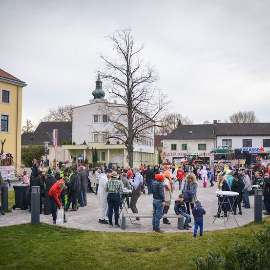 Eine große Menschenmenge in Kostümen versammelt sich auf einem Freiluftplatz mit Gebäuden im Hintergrund, unter einem bewölkten Himmel.