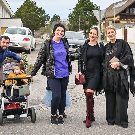 Eine vierköpfige Familie, einschließlich eines Babys in einem Kinderwagen, steht auf einer Straße mit geparkten Autos und Gebäuden im Hintergrund.