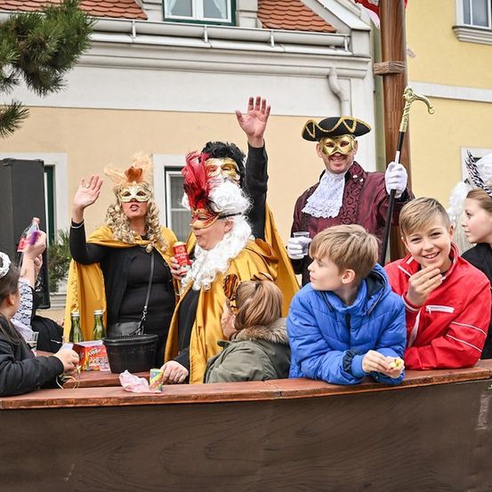 Eine Gruppe von Menschen in Kostümen, einschließlich Masken, sitzt in einem Boot und scheint eine festliche Veranstaltung zu genießen. Ein Mann in einem Piratenkostüm hält eine Dose und einen Stab.