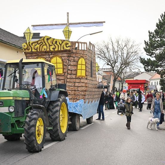 Ein dekorierter Traktor mit einer burgartigen Struktur auf dem Rücken fährt während einer Parade die Straße entlang. Menschen gehen auf dem Bürgersteig, und einige stehen in der Nähe eines roten Lastwagens. Bäume und Gebäude sind im Hintergrund zu sehen.