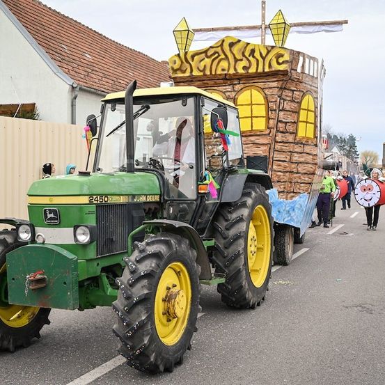 Ein dekorierter Traktor mit einer burgartigen Struktur auf dem Rücken fährt die Straße entlang. Menschen in Kostümen gehen dahinter.