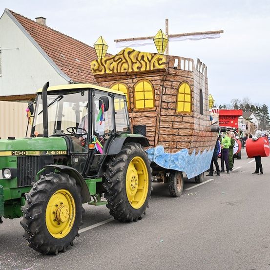 Ein John Deere Traktor zieht einen geschmückten Nachbau eines Schiffes während einer Parade, mit Menschen in Kostümen dahinter.