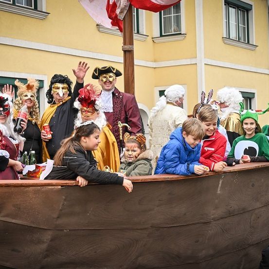 Eine Gruppe von Menschen in Kostümen, einschließlich Masken und Gesichtsbemalung, steht auf einem hölzernen Boot, möglicherweise für einen Umzug. Sie lächeln und winken. Hinter ihnen steht ein Gebäude mit Fenstern und einer Flagge.