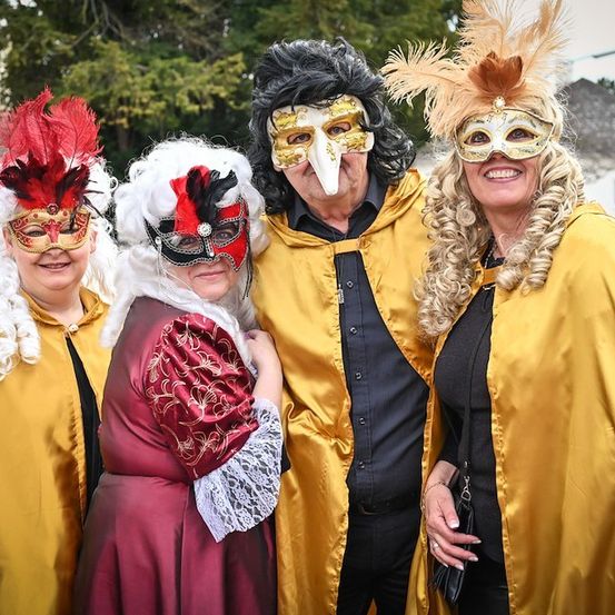Vier Menschen in farbenfrohen Kostümen und Masken stehen eng beieinander und lächeln für die Kamera. Sie stehen vor einem Gebäude mit Bäumen im Hintergrund.