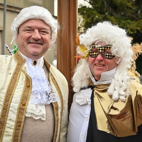 Zwei Männer in aufwendigen historischen Kostümen, einer mit weißer Perücke und goldenem Umhang, der andere mit weißer Perücke und schwarzer Maske, beide lächeln in die Kamera.