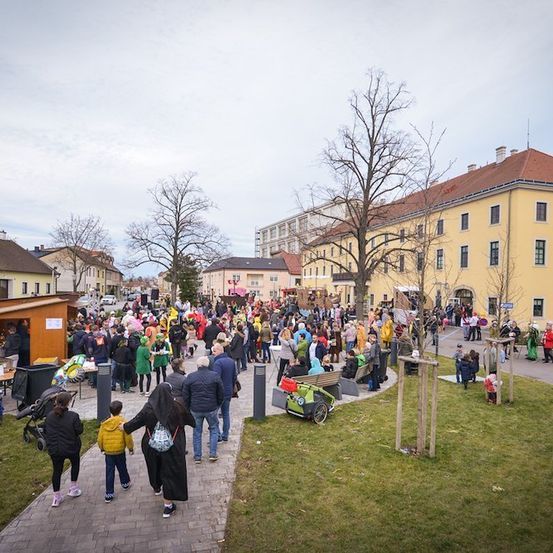 Eine große Menschenmenge, darunter Familien und Kinder, versammelt sich in einem Außenbereich mit Gebäuden und Bäumen, möglicherweise für eine Veranstaltung. Einige tragen Kostüme.