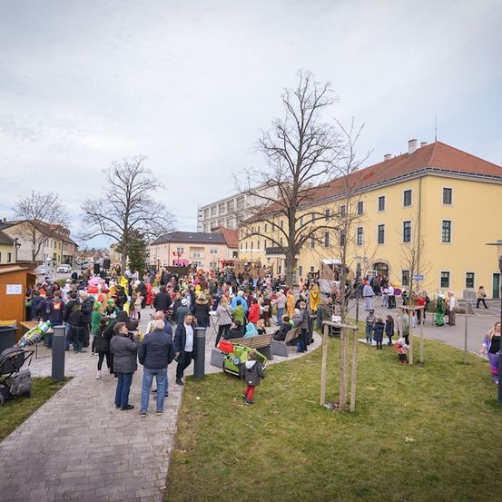 Eine große Menschenmenge in bunten Kostümen versammelt sich auf einem Stadtplatz mit gelben Gebäuden und Bäumen. Einige gehen, während andere auf Bänken sitzen. Ein Kinderwagen und ein Mülleimer sind links.