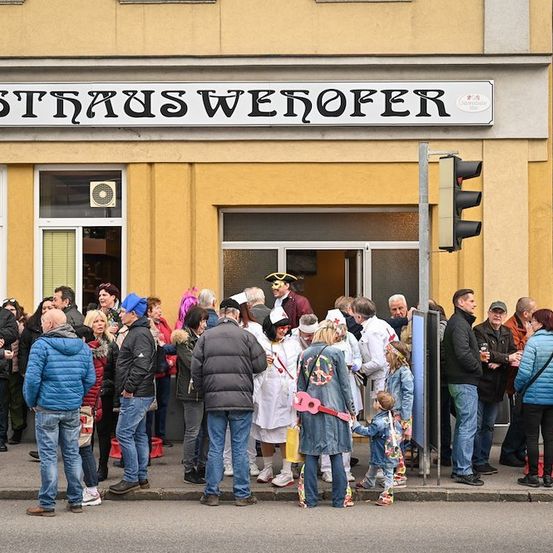 Eine Menschenmenge in verschiedenen Kostümen, darunter einige in weißen Laborkittel, versammelt sich vor einem Gebäude mit dem Schild 'Strauss Wenofer'. Einige halten Getränke in der Hand und unterhalten sich, während andere in der Nähe einer Ampel stehen.