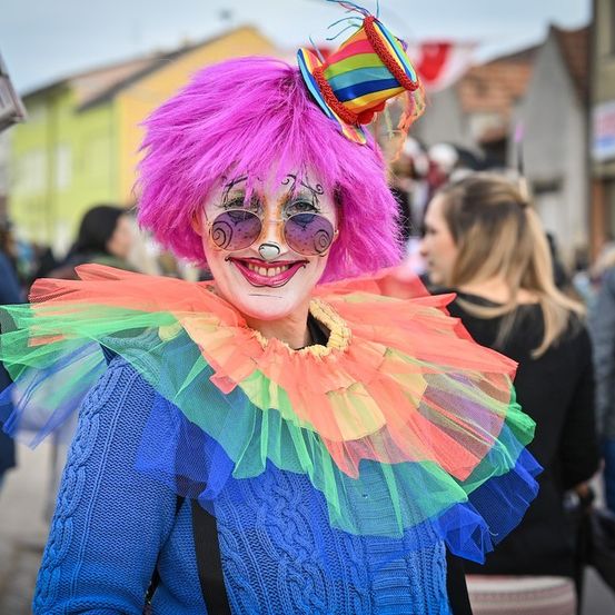 Eine Person, die als Clown verkleidet ist, mit rosa Haaren, bunten Tüllröcken und Gesichtsbemalung, steht lächelnd auf der Straße. Im Hintergrund sind verschwommen andere Menschen und Gebäude zu sehen.