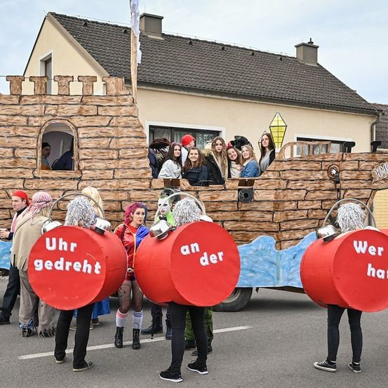 Eine Gruppe von Menschen in Kostümen steht vor einem Piratenschiff-Float bei einer Parade, einer hält eine rote Bombenattrappe mit der Aufschrift 'Uhr gedreh'.