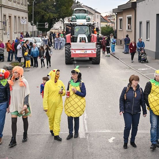 Ein Straßenumzug mit Menschen in Kostümen, darunter ein Clown und Ananas-Outfits, geht vor einem Traktor und einer Menge von Zuschauern.