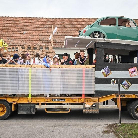 Ein gelber LKW mit einem grünen Auto auf dem Dach und Leuten, die darauf fahren. Der LKW ist mit Aufklebern und einem weißen Tuch dekoriert. Die Leute tragen Kostüme und einige halten Getränke. Der LKW ist am Straßenrand geparkt.