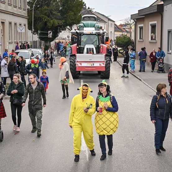 Eine Straße voller Menschen in Kostümen, darunter eine Frau in einer Ananas-Kostüm und ein Mann in einem Clown-Kostüm, gehen an einem roten Traktor vorbei.