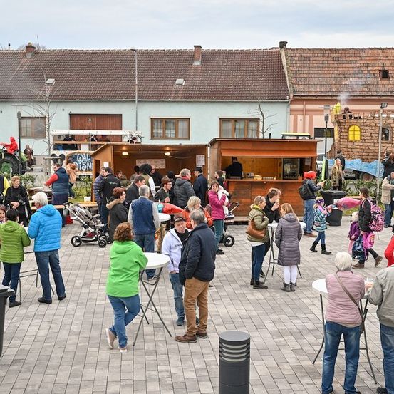 Eine Gruppe von Menschen versammelt sich auf einem Stadtplatz, einige stehen um Tische herum und andere gehen herum. Im Hintergrund befinden sich Essensstände und ein Traktor.