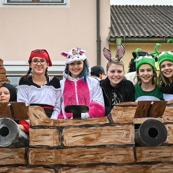 Eine Gruppe junger Menschen in Kostümen posiert für ein Foto vor einer Holzstruktur. Sie lächeln und scheinen Spaß zu haben.