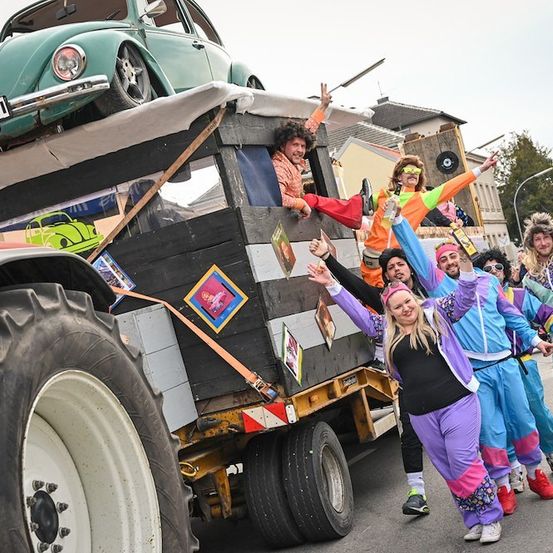 Eine Gruppe von Menschen in bunten Disco-Outfits steht auf einem Lastwagen mit einem grünen VW-Käfer obenauf und posiert für ein Foto.