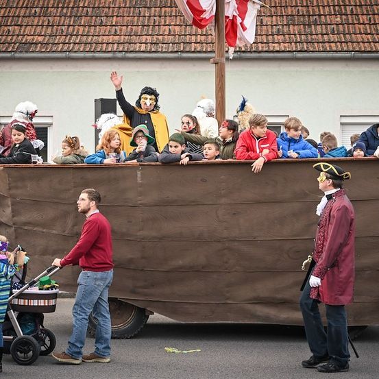 Eine Gruppe von Menschen in Kostümen auf einem Wagen, mit einem Mann in einem roten Mantel, der an der Seite steht. Ein Mann schiebt einen Kinderwagen vor dem Wagen.