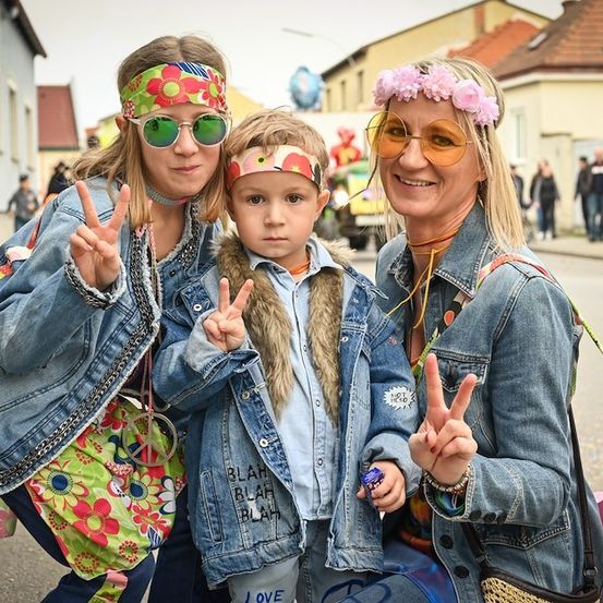 Drei Menschen, einschließlich eines jungen Jungen, posieren für ein Foto auf einer Straße. Die beiden Frauen tragen Stirnbänder und Jeansjacken, während der Junge eine mit Fell gefütterte Jacke trägt. Sie machen alle Friedenszeichen.