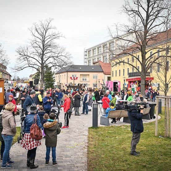 Eine Menschenmenge versammelt sich auf einem Stadtplatz, einige stehen und andere sitzen auf Bänken. Ein Fotograf hält die Szene fest, und ein rosafarbener Imbisswagen ist geparkt. Gebäude mit gelben Wänden säumen die Straße.