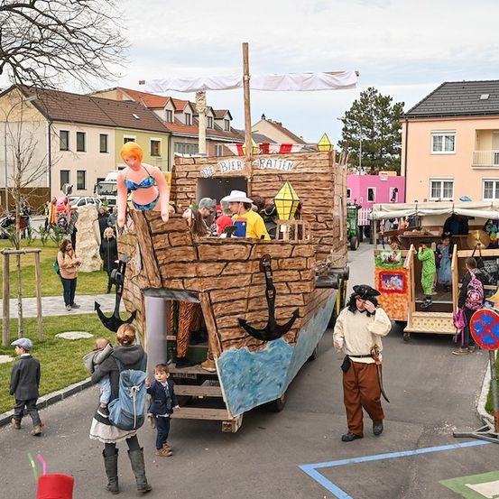 Ein pirat-thematischer Wagen steht auf einer Straße, um ihn herum sind Leute. Eine Frau in einem Bikini ist auf dem Wagen. Ein Mann in einem Piratenhut geht in der Nähe.