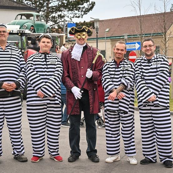 Fünf Männer in gestreiften Gefängnisuniformen, einer trägt einen Piratenhut und eine Maske, stehen auf einer Straße.