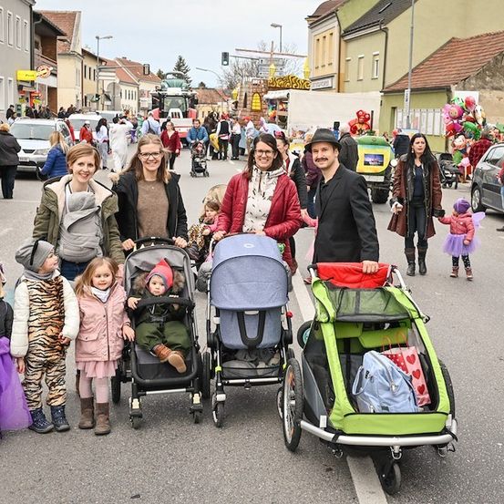 Eine Gruppe von Familien mit kleinen Kindern steht mitten auf einer Straße während eines Umzugs, mit Kinderwagen und Luftballons im Hintergrund.