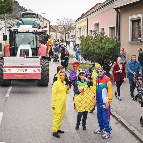 Eine Gruppe von Menschen in bunten Kostümen geht auf der Straße, mit einem Traktor im Hintergrund.