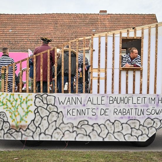 Eine Gruppe von Menschen steht auf einem Wagen mit einer Holzstruktur und Graffiti. Ein Mann schaut aus einem Fenster. Hinter dem Wagen befindet sich ein Haus mit einer rosa Wand.