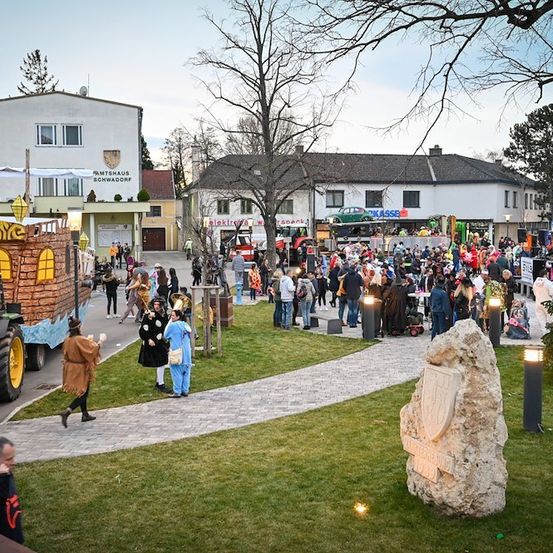 Eine lebhafte Straßenszene mit einem festlichen Umzugswagen, einer Menschenmenge und einem Traktor im Vordergrund. Gebäude säumen die Straße, und es gibt Straßenlaternen und Bäume im Hintergrund.
