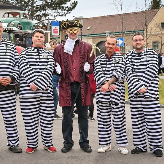 Fünf Männer in gestreiften Kostümen, darunter einer in einem Piratenhut, stehen in einer Reihe auf einer Straße mit einem Gebäude und geparkten Autos im Hintergrund.