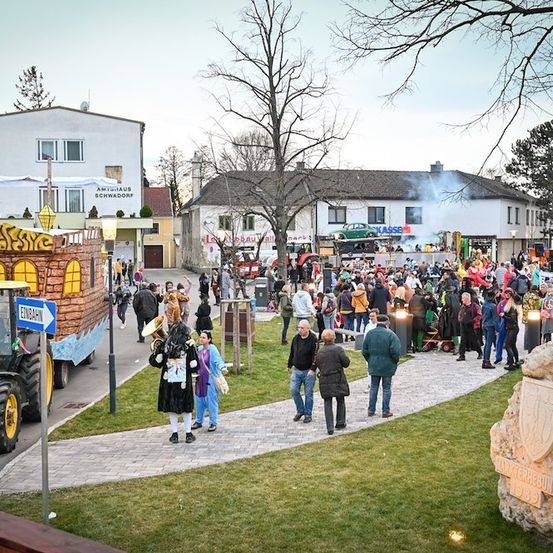 Eine lebhafte Straßenszene mit einem geschmückten Festwagen, einer Marschkapelle und einer Menschenmenge, die auf einem gepflasterten Weg geht. Im Hintergrund sind Gebäude, Bäume und ein Traktor zu sehen.
