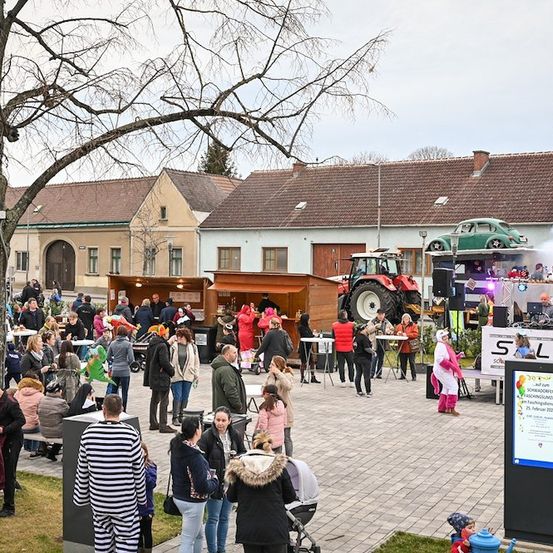 Ein lebhaftes Outdoor-Event mit einer Menschenmenge, Essensständen, einem Traktor und einem Auto zur Schau. Die Umgebung umfasst Gebäude, Bäume und eine Bühne mit einem DJ.