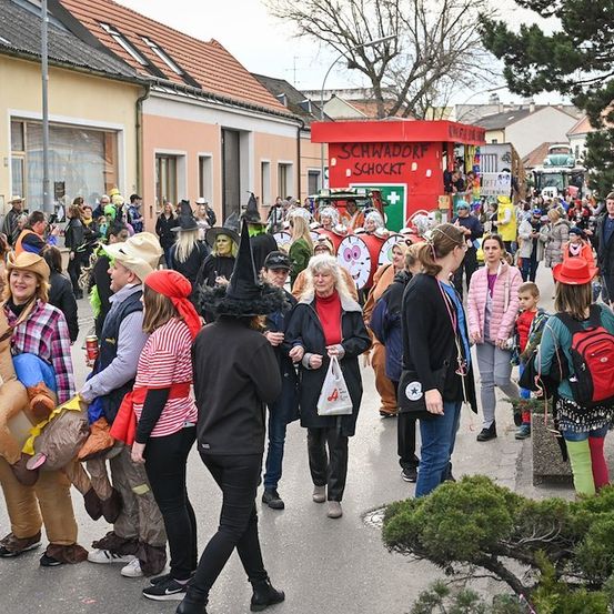 Eine Menschenmenge in verschiedenen Kostümen, darunter Hexen und Tiere, geht während einer Veranstaltung im Freien auf einer Straße. Im Hintergrund ist ein roter Stand mit der Aufschrift 'SCHWADRORF SCHOCKT' zu sehen.