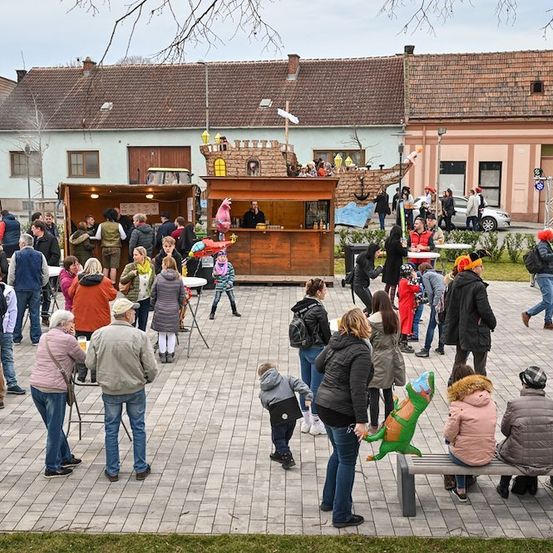 Eine Menschenmenge versammelt sich im Freien, einige sitzen auf Bänken und andere stehen. Es gibt Essensstände und eine Piratenschiffdekoration. Ein Mann steht hinter einer Theke an einem der Stände. In der Ferne befinden sich Gebäude und ein Auto.