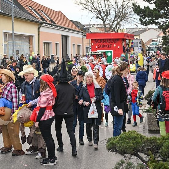 Eine Menschenmenge in Kostümen, die eine Straße entlanggeht, einige in aufblasbaren Kostümen, mit Gebäuden und einer roten Bude im Hintergrund.