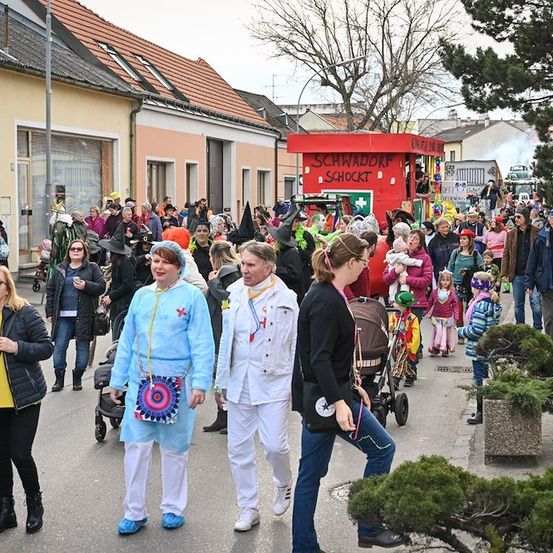 Eine Menschenmenge in verschiedenen Kostümen, darunter einige in medizinischen Gewändern, geht während eines Umzugs auf einer Straße. Im Hintergrund sind Gebäude und ein roter Stand zu sehen.