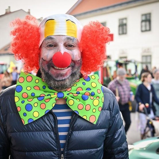 Ein Mann, der als Clown verkleidet ist, mit roten Locken, einer herzförmigen roten Nase und einer gepunkteten Schleife steht vor einem Gebäude.