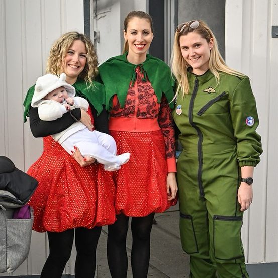 Drei Frauen in festlichen Kostümen, eine hält ein Baby, stehen vor einem Gebäude. Die Frau links trägt ein rotes Kleid, die mittlere ein rotes Kleid mit grünem Umhang und die rechte ein grünes Overall.