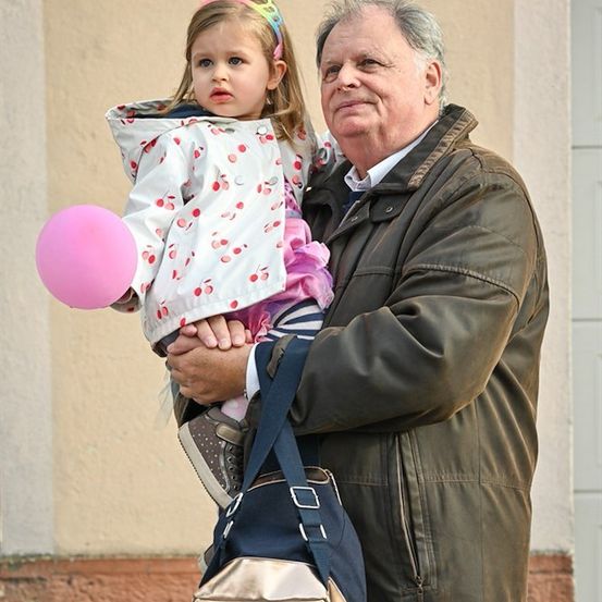 Ein älterer Mann hält ein kleines Mädchen mit einem rosa Luftballon vor einem Gebäude.