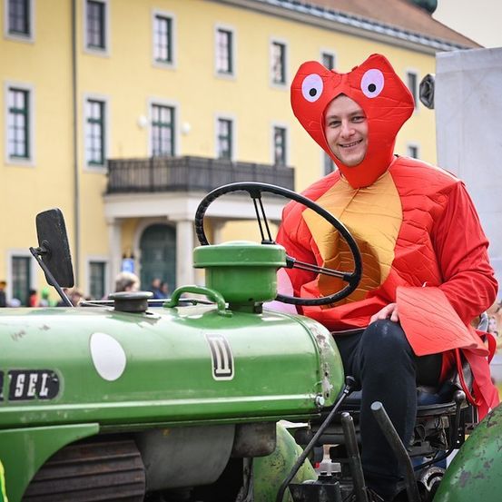 Eine Person in einem roten Kostüm mit einer Fischkopfmaske sitzt auf einem grünen Traktor vor einem gelben Gebäude.