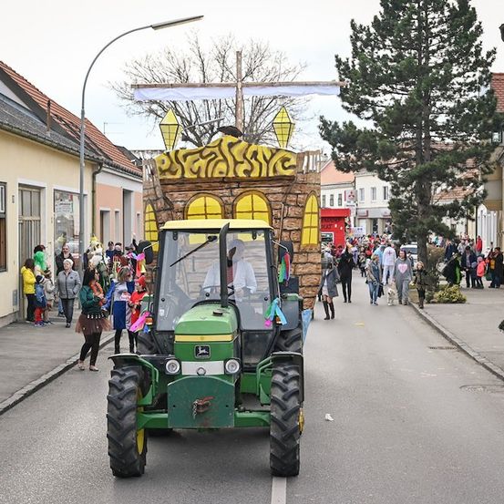 Ein geschmückter Traktor fährt während einer Parade durch eine Straße, mit Menschen, die auf dem Bürgersteig gehen, und Gebäuden auf beiden Seiten.