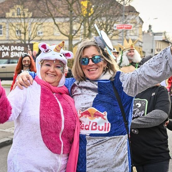 Zwei Frauen in Kostümen, eine als Einhorn verkleidet, posieren für ein Foto auf der Straße mit einer Menge und Gebäuden im Hintergrund.