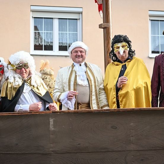 Fünf Personen in historischen Kostümen stehen auf einer Holzplattform und lächeln für die Kamera. Hinter ihnen steht ein Gebäude mit zwei Fenstern und einem Fahnenmast.