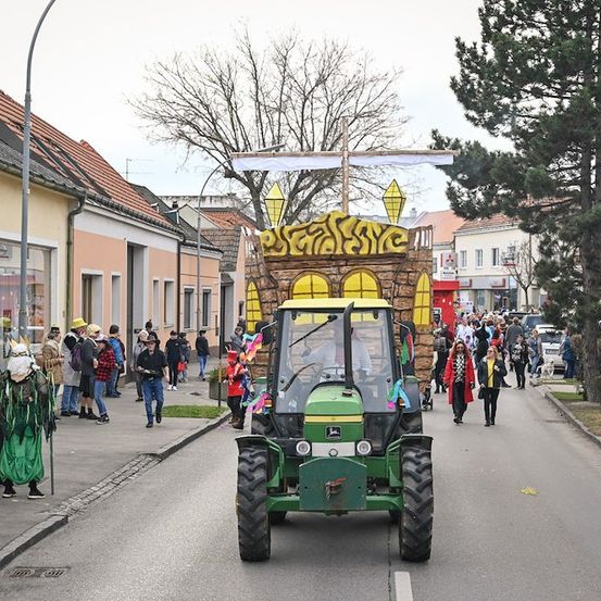 Ein geschmückter Traktor mit einer burgartigen Struktur auf dem Rücken fährt während einer Parade die Straße hinunter, wobei Menschen in Kostümen am Bürgersteig zusehen.