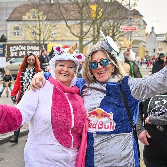 Zwei Frauen in Kostümen, eine in einem rosa Einhorn-Overall und die andere in einem blauen Red Bull-Outfit, posieren für ein Foto auf einer Straße mit anderen Leuten und Gebäuden im Hintergrund.